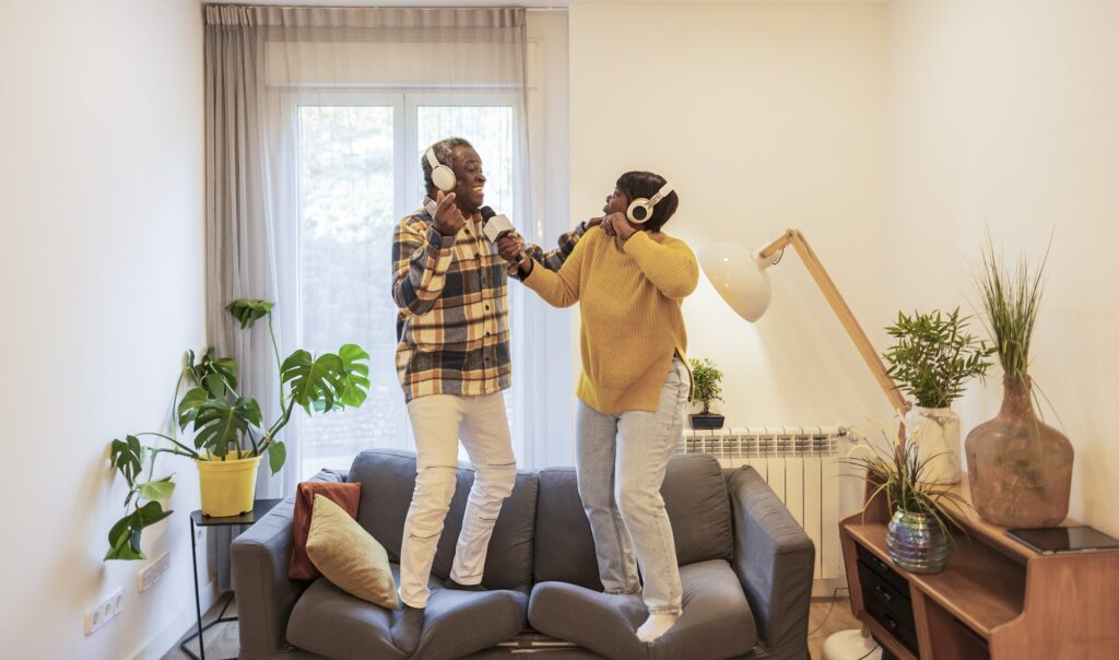 Happy active senior couple play and dance and sing on the couch at home in headphones listening